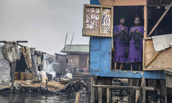 Hai người phụ nữ mặc áo màu tím đang đứng ở bậc cửa của một quán tóc và làm đẹp ở khu vực ven biển Makoko ở Lagos, Nigeria. Họ thường rất bận rộn vào ngày Chủ nhật, khi những người phụ nữ địa phương chuẩn bị đi lễ nhà thờ hay các nghi lễ khác. Khu ổ chuột Makoko là một cộng đồng bị đe dọa đuổi nhà, chứa hàng chục nghìn người sống trong các nhà nổi trên dòng nước bị ô nhiễm của đầm Lagos. Ảnh: Petrut Calinescu