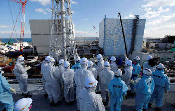 Thành viên của các phương tiện truyền thông mặc quần áo bảo hộ và đeo khẩu trang được các nhân viên của Công ty điện lực Tokyo (TEPCO) (mặc đồ màu xanh lá cây) hướng dẫn ở trước 2 tòa nhà lò phản ứng số 1 (bên trái) và số 2 tại nhà máy điện hạt nhân Fukushima Daiichi của TEPCO bị sóng thần tàn phá. Ảnh: REUTERS / Toru Hanai
