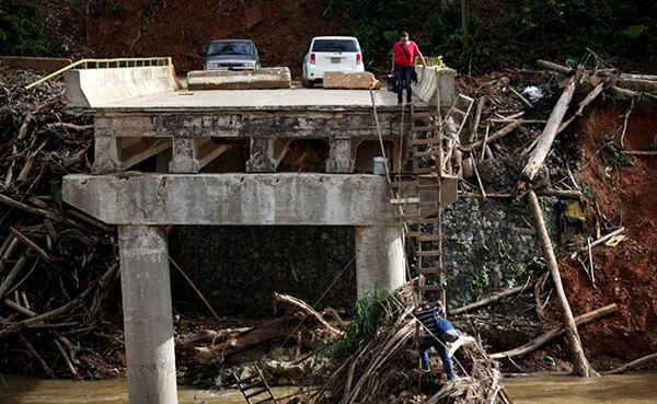 Người phụ nữ nhìn xuống người chồng của cô đang trèo thang xuống dưới một cây cầu bị phá hủy sau khi bão Maria tấn công Utuado, Puerto Rico hồi tháng 9/2017. Ảnh chụp ngày 9/11/2017 của tác giả Alvin Baez